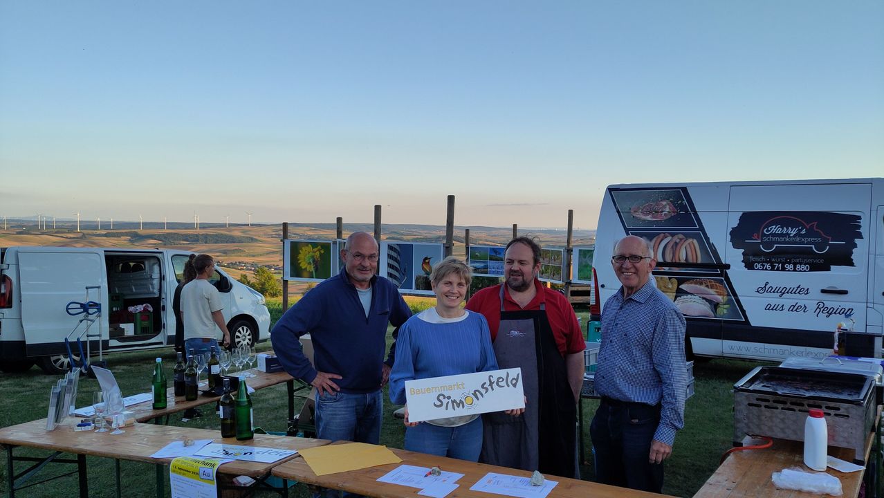 Vier Personen stehen lächelnd an einem Tisch. Eine Frau hält ein Schild mit der Aufschrift 'Baumarkt Simonsfeld'. Hinter ihnen stehen mehrere Bilderrahmen auf Stangen, ein Van und ein Auto. In der Ferne befinden sich Hügel und Windmühlen.