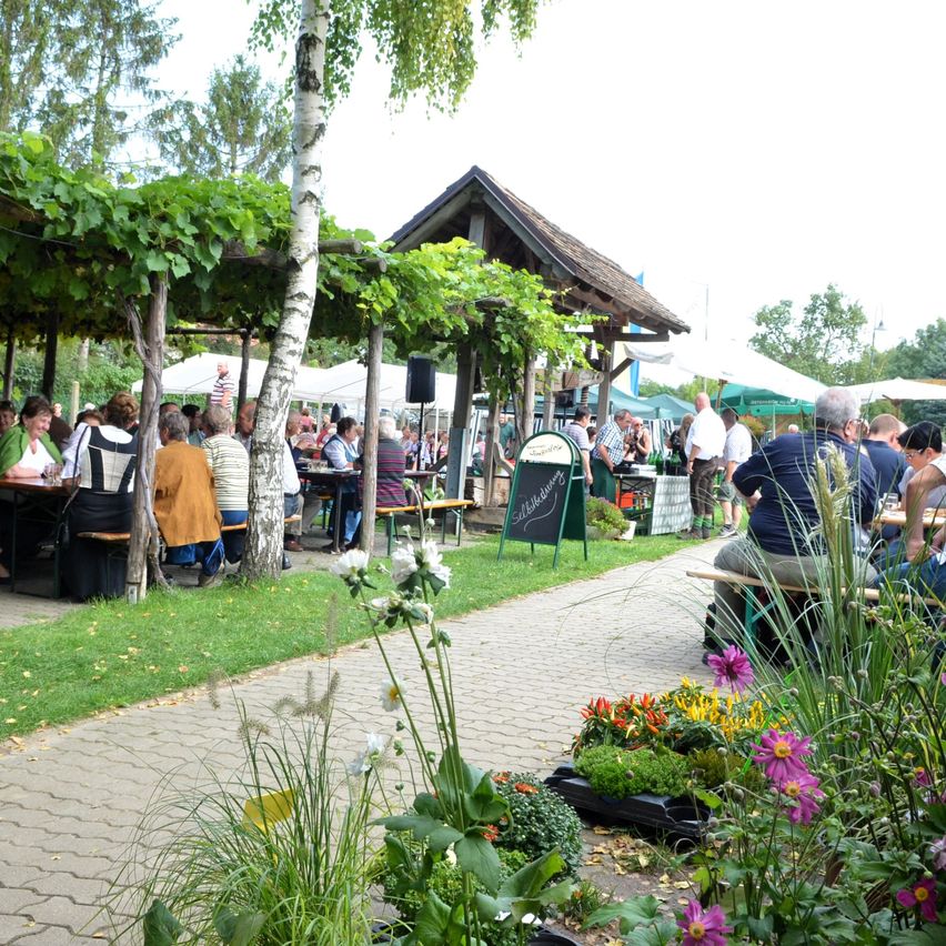 Ein Gartenrestaurant mit Menschen, die an Tischen unter einer Pergola sitzen. In der Nähe befinden sich weiße Zelte und eine Tafel mit Schrift. Das Gebiet ist von Bäumen und Blumen umgeben.