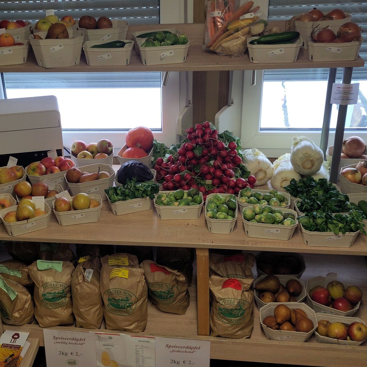 Ein Holzregal zeigt eine Vielzahl frischer Produkte, darunter Äpfel, Karotten, Zwiebeln und verschiedene Grüngemüse in weißen Körben. Säcke mit Produkten sind auch auf dem unteren Regal sichtbar.
