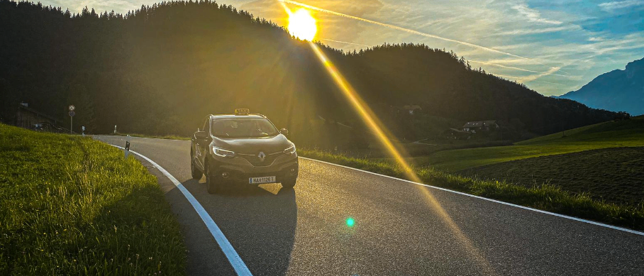 Ein schwarzer Renault SUV fährt auf einer kurvenreichen Straße, mit einer strahlenden Sonne hinter ihm. Die Straße ist von saftigem grünem Gras umgeben und in der Ferne erheben sich Berge.