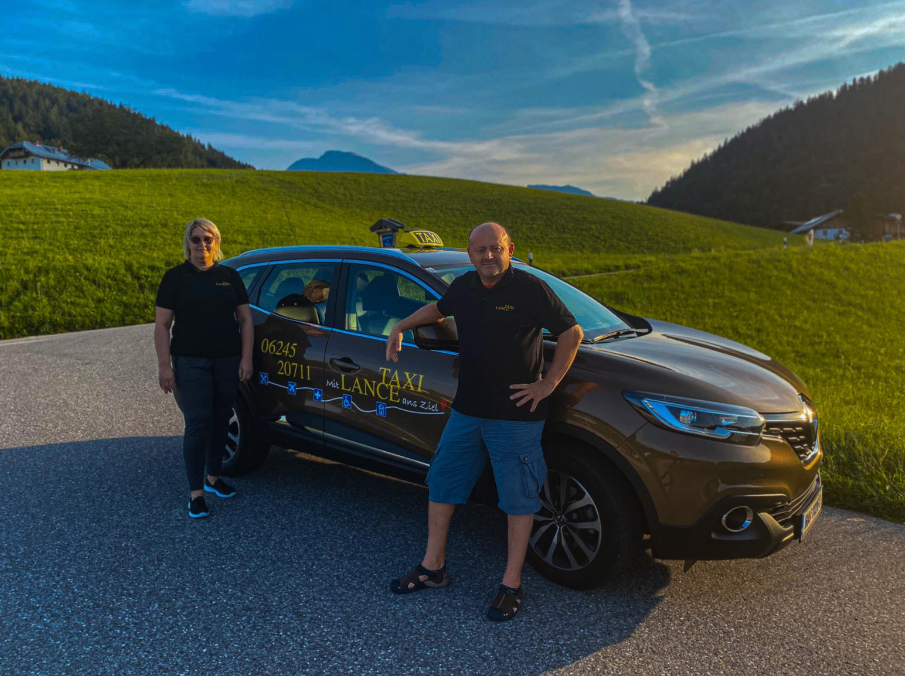 Zwei Personen stehen vor einem Taxi mit einem Taxischild auf dem Dach. Sie tragen beide schwarze T-Shirts. Das Taxi hat ein Nummernschild, das 06245 20711 liest.