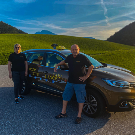 Zwei Personen stehen vor einem Taxi mit einem Taxischild auf dem Dach. Sie tragen beide schwarze T-Shirts. Das Taxi hat ein Nummernschild, das 06245 20711 liest.