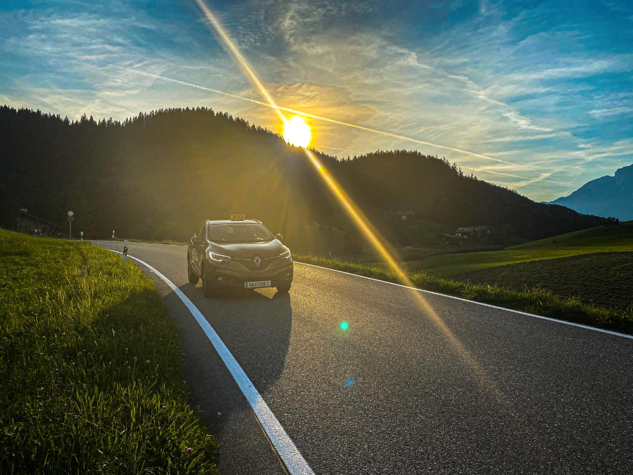 Ein Auto fährt auf einer Straße mit einem Bergrücken und Bäumen auf der Seite. Die Sonne geht hinter dem Berg unter.