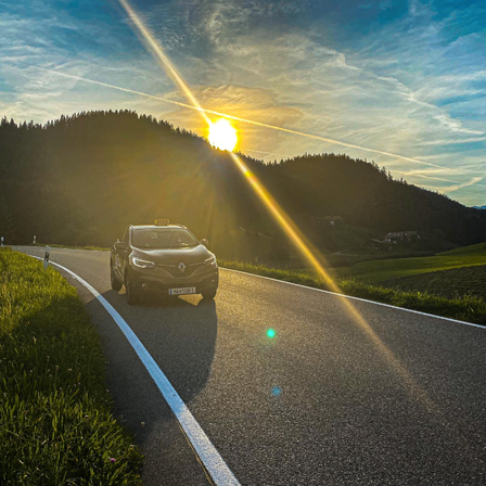 Ein Auto fährt auf einer Straße mit einem Bergrücken und Bäumen auf der Seite. Die Sonne geht hinter dem Berg unter.