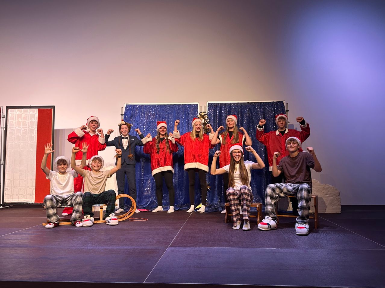 Eine Gruppe von Menschen in Weihnachtskostümen steht auf der Bühne. Sie posieren für ein Foto mit erhobenen Händen. Dahinter befindet sich ein blauer Hintergrund mit einem weißen Vorhang. Es gibt zwei Schlitten auf der Bühne.