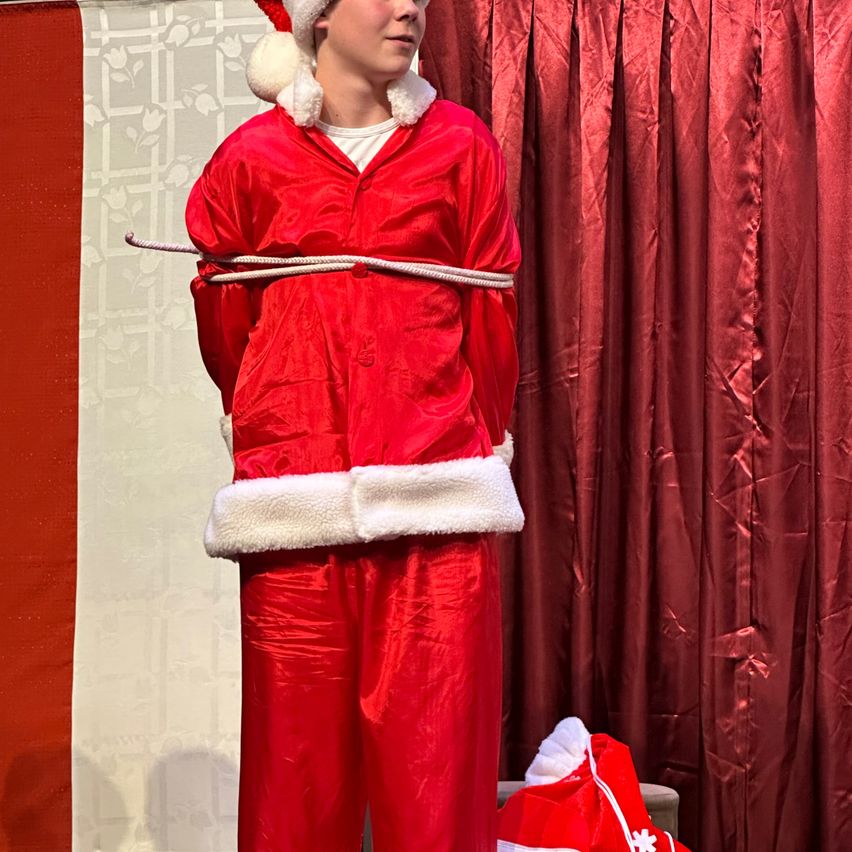 Ein junger Mann in einem roten Weihnachtsmannkostüm mit weißem Pelzbesatz steht vor einem roten Vorhang. Er trägt einen Weihnachtsmannhut und hat einen Seil um die Taille gebunden.