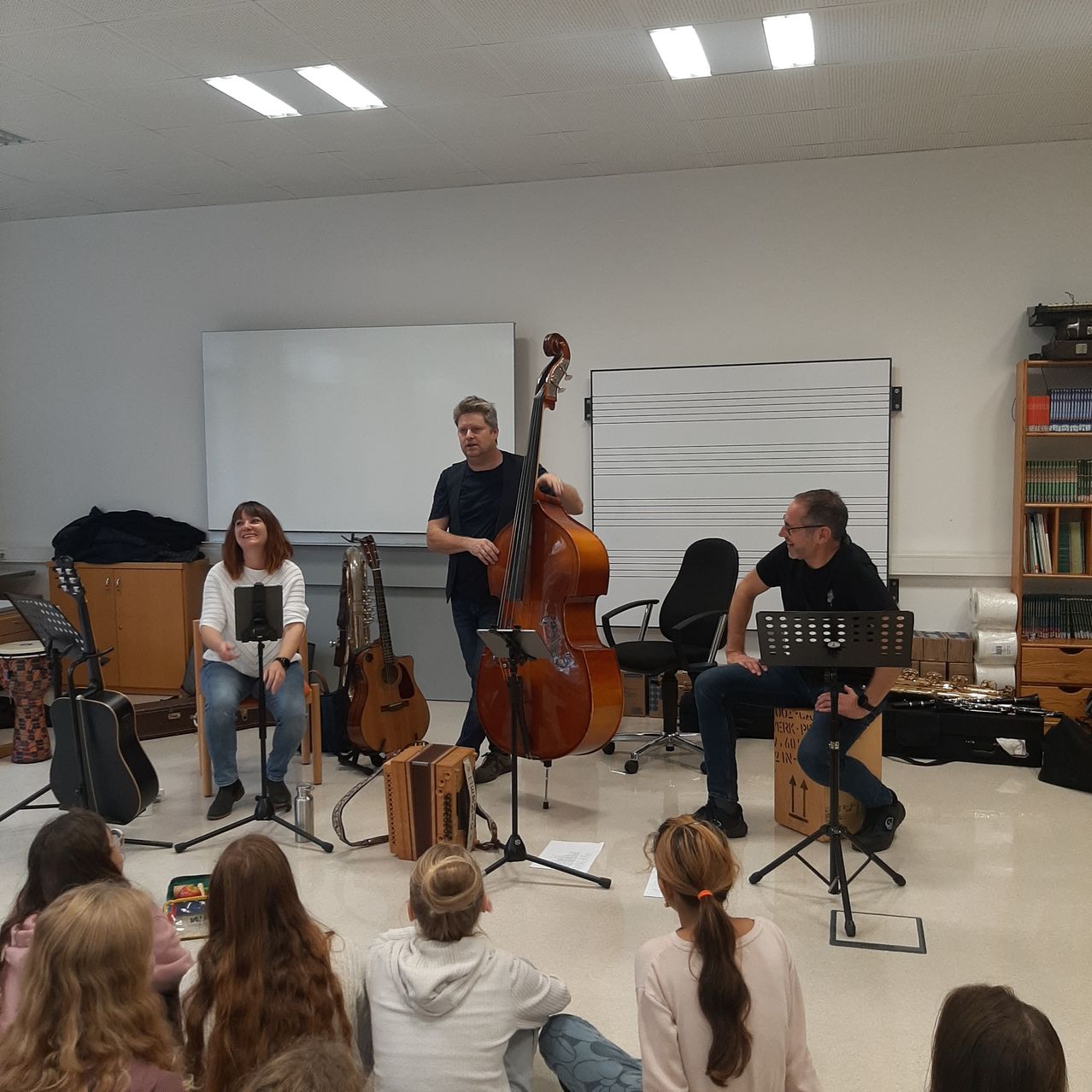 Drei Musiker, einer spielt Cello, einer Gitarre und einer Akkordeon, spielen für eine Gruppe von Kindern in einem Klassenzimmer.