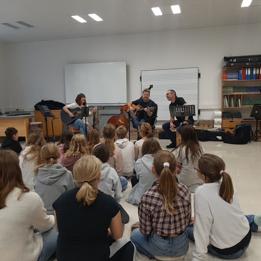 Eine Gruppe von Menschen sitzt auf dem Boden in einem Klassenzimmer und lauscht einer Gitarren- und Cello-Darbietung von zwei Männern.