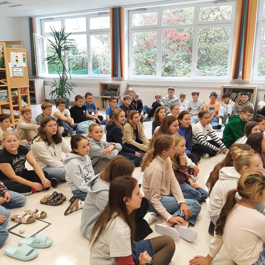 Ein Klassenzimmer voller Schüler, die auf dem Boden sitzen, einige tragen Hoodies, Sneakers und Sandalen. Sie sind versammelt, wahrscheinlich um einem Lehrer zuzuhören. Es gibt Topfpflanzen und Bücherregale im Raum.