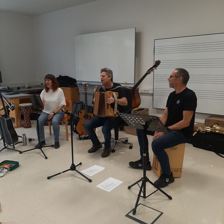 Drei Musiker sitzen in einem Raum mit Instrumenten. Eine Frau steht, während zwei Männer Gitarre und Akkordeon spielen. Eine Tafel und ein Notenblatt sind hinter ihnen.