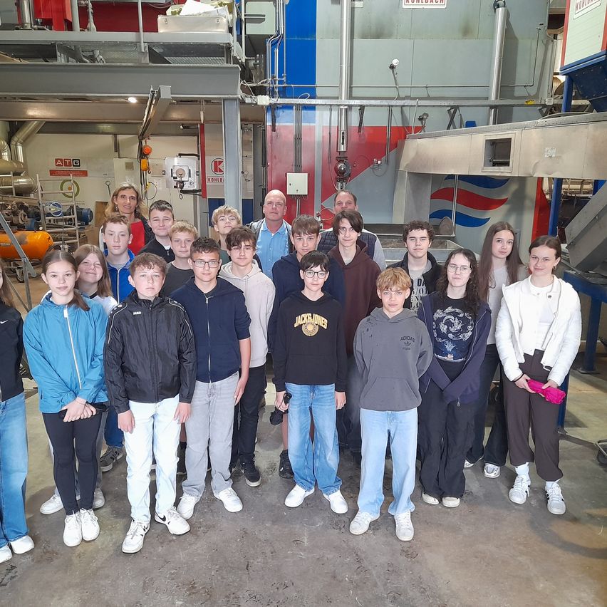 A group of students and two adults are posing for a picture in an industrial area. Behind them are large metal structures and machinery.