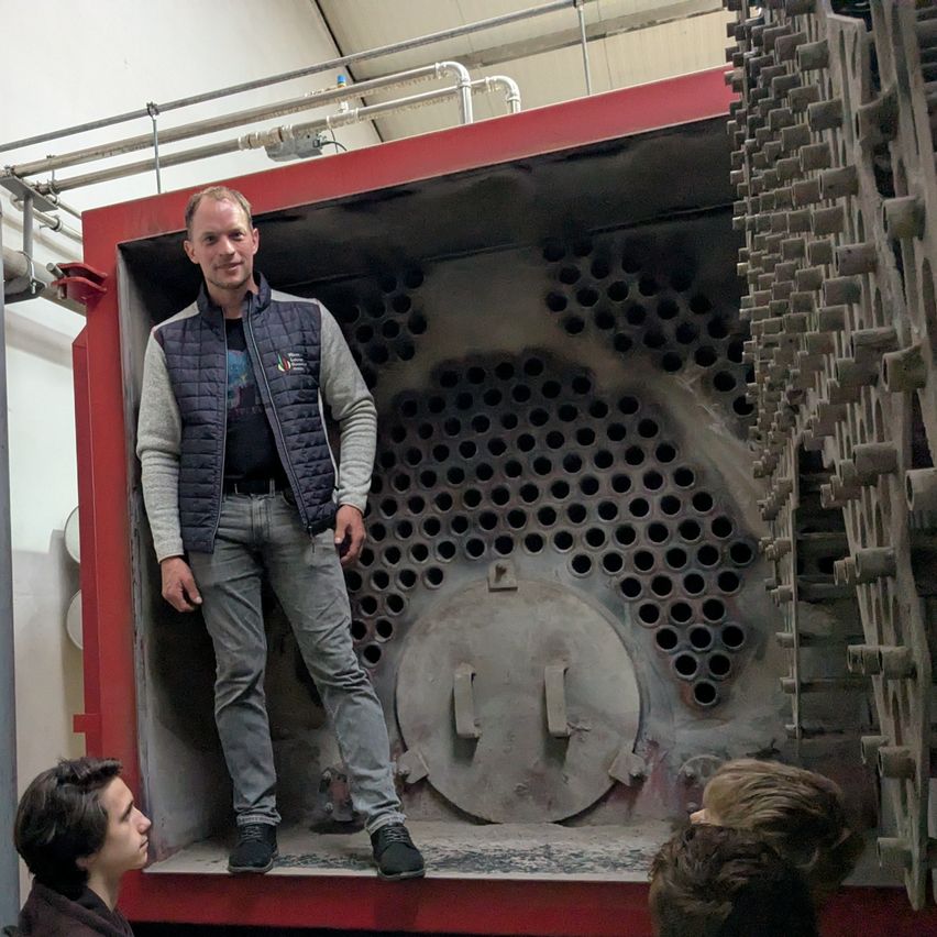 A man stands inside a large red machine with a holed circular cover. He wears a vest and jeans. Behind him, two people watch curiously.