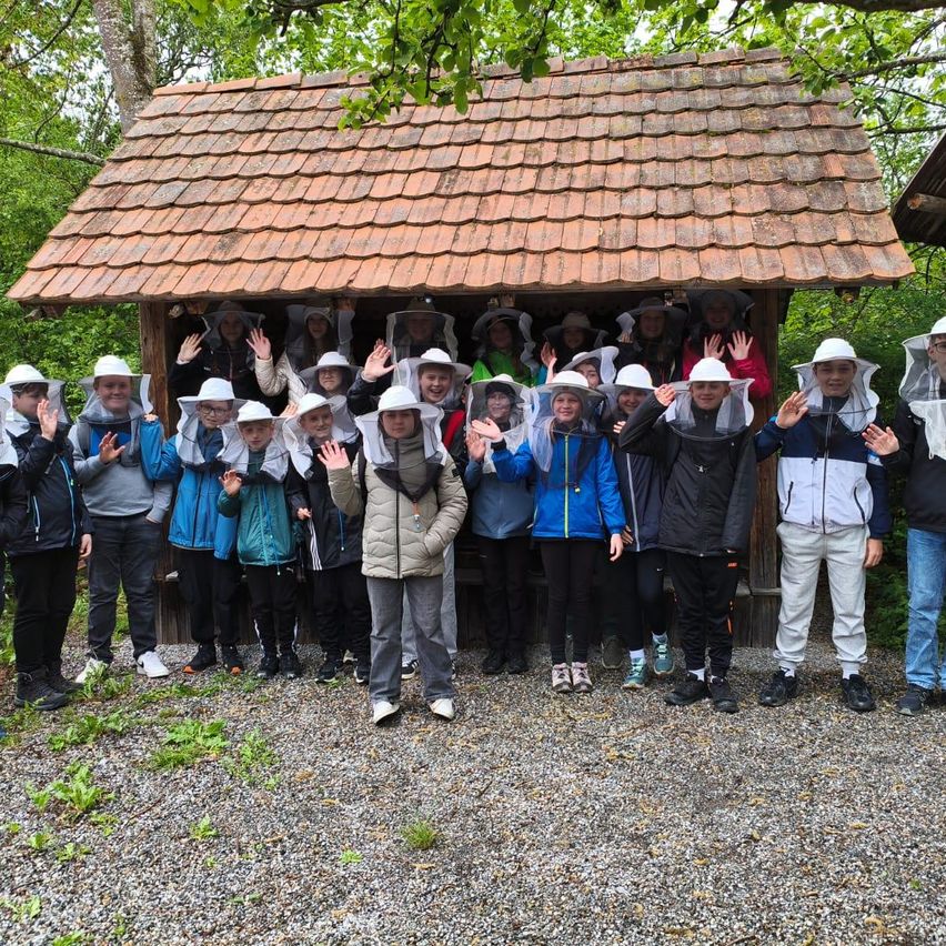 A group of people wearing beekeeper gear stands in front of a small wooden house with a tiled roof, surrounded by trees. They appear to be posing for a photo.