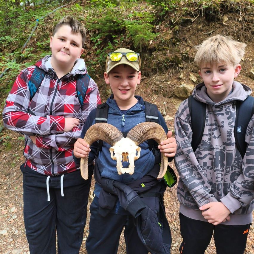 Three boys stand in a forest, smiling. One holds a ram skull. The boy on the left wears a plaid jacket, the middle one a cap and goggles, and the right one a gray hoodie.