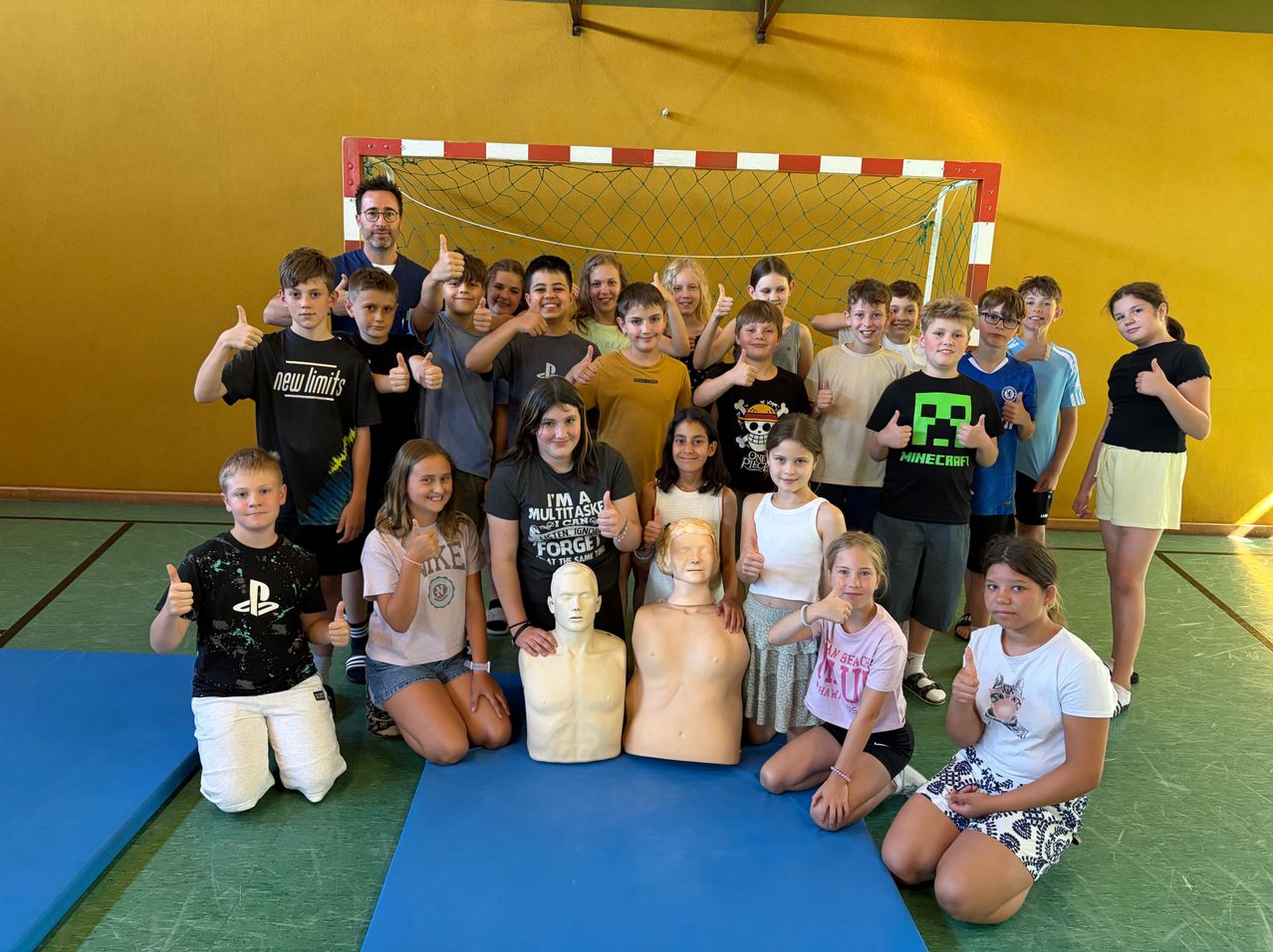 Gruppenfoto von Kindern und zwei Erwachsenen, die mit erhobenen Daumen in einer Turnhalle posieren. Zwei Puppen liegen auf der blauen Matte vor ihnen. Ein Fußballtor ist im Hintergrund.