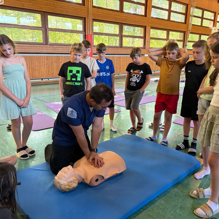 Ein Mann zeigt einer Gruppe von Kindern auf einer blauen Matte, wie man Herzdruckmassage durchführt. Die Kinder stehen um den Mann herum, einige mit den Händen am Kopf.