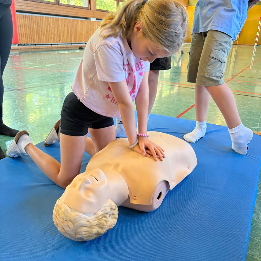 Ein junges Mädchen übt in einer Turnhalle die Herz-Lungen-Wiederbelebung an einem Mannequin. Sie kniet über dem Dummies, das auf einem blauen Matten liegt, und führt Brustkompressionen durch. Eine andere Person steht in der Nähe und beobachtet.