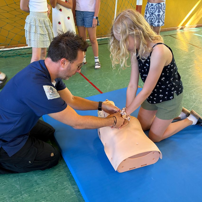 Ein Mann mit Brille und eine Frau üben CPR an einem Dummy in einer Turnhalle. Andere Leute beobachten.