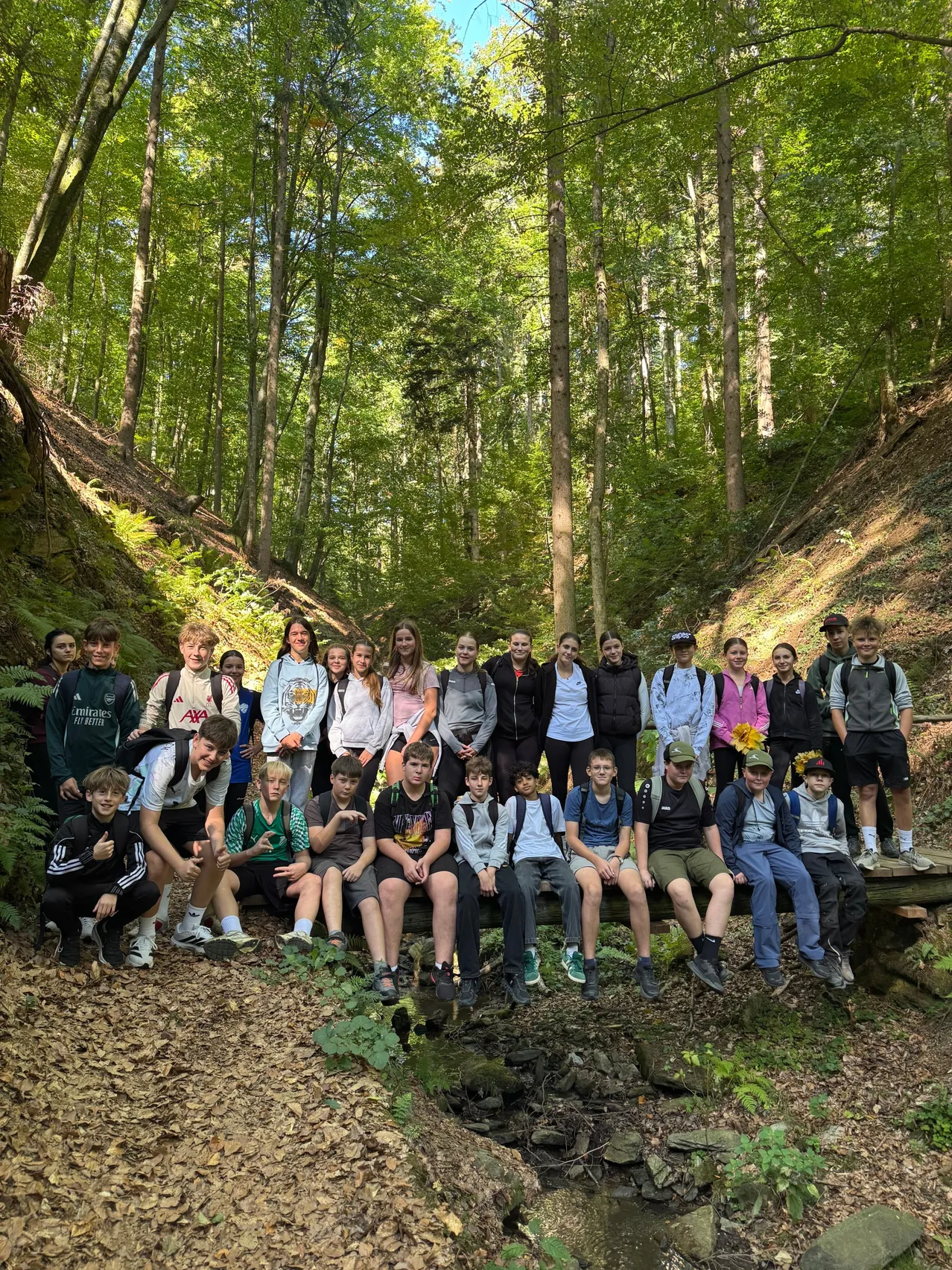 Eine Gruppe junger Menschen posiert für ein Foto im Wald. Sie sitzen auf einer Holzbrücke und einige stehen an der Seite.
