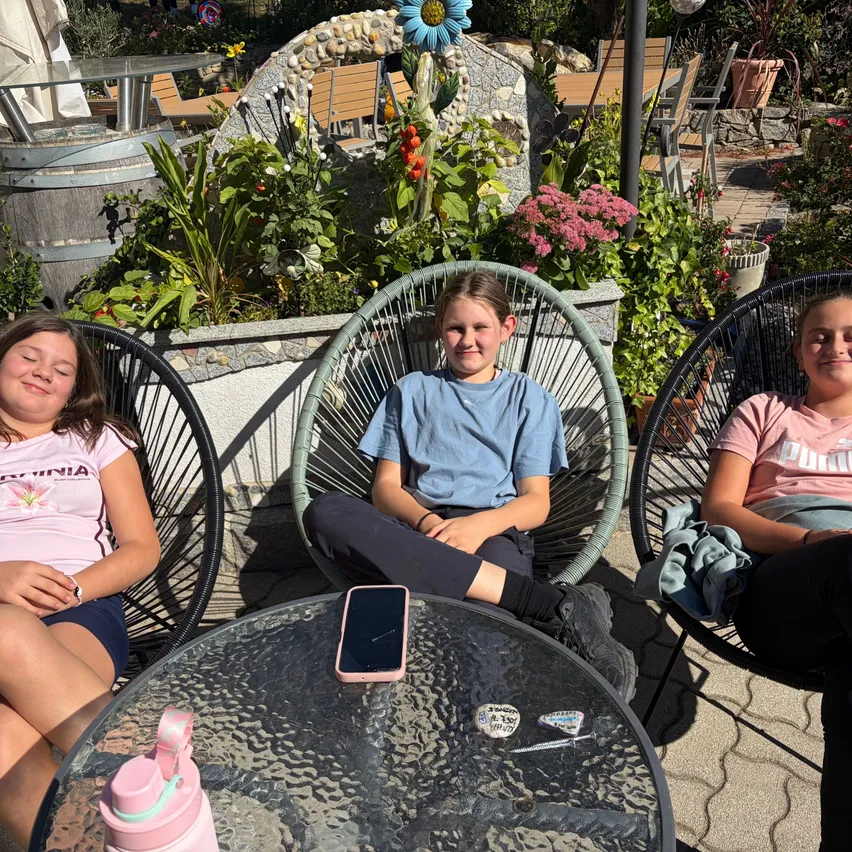 Drei junge Mädchen sitzen auf Stühlen in einem Garten. Ein Mädchen trägt ein rosa Shirt mit Blumendesign, ein anderes trägt ein blaues Shirt und das dritte trägt ein rosa Shirt mit Logo. Sie sitzen um einen Tisch herum, auf dem ein Handy und eine rosa Wasserflasche stehen.