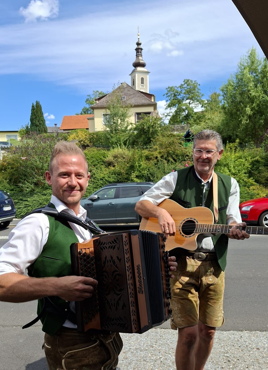 Zwei Männer lächeln und posieren für ein Foto in einer Stadt. Einer spielt Akkordeon und der andere Gitarre. Hinter ihnen steht eine Kirche, und geparkte Autos sind im Vordergrund.