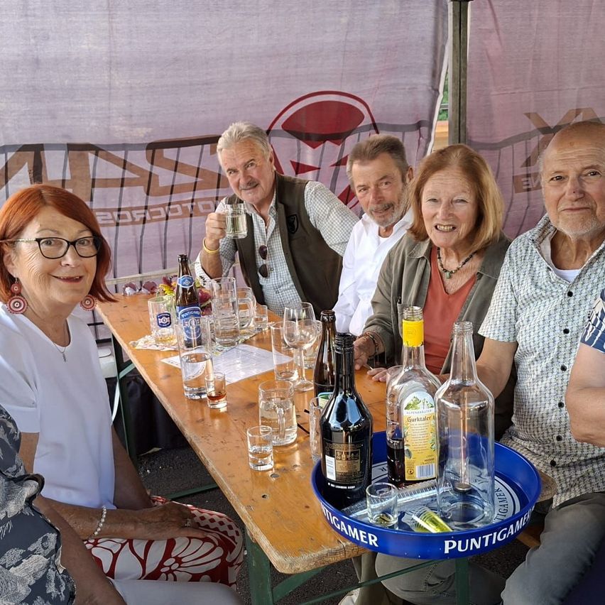 Fünf Personen, zwei Frauen und drei Männer, sitzen um einen Holztisch herum, der mit Gläsern und Flaschen gefüllt ist. Der Tisch ist mit einem blauen Tablett geschmückt, auf dem 'Puntigamer' steht. Die Umgebung scheint eine gesellige Zusammenkunft mit guter Laune zu sein.