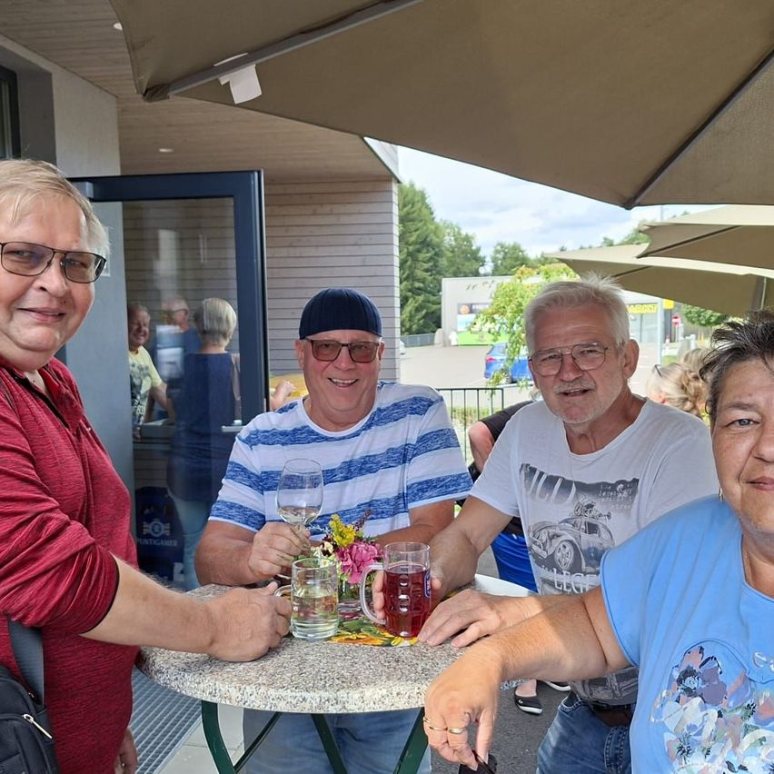 Vier Erwachsene lächeln und posieren für ein Foto, sitzend um einen kleinen Tisch mit Getränken, unter großen Sonnenschirmen vor einem Gebäude.