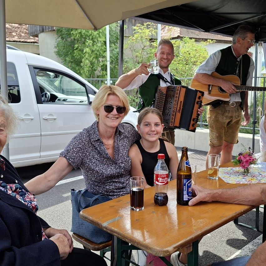 Eine Gruppe von Menschen, darunter zwei Frauen, ein junges Mädchen und zwei Männer, sitzen um einen Tisch unter einem Regenschirm. Die Männer spielen Musikinstrumente. Ein Van ist hinter ihnen geparkt.