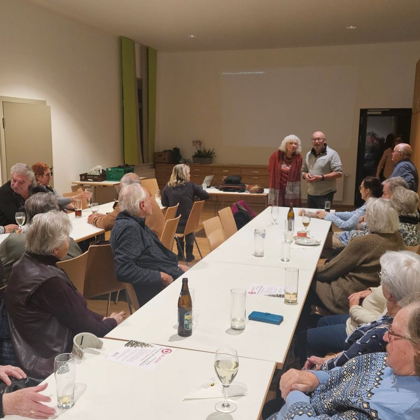 Mehrere ältere Menschen sitzen an Tischen in einem Raum, einige stehen. Ein Mann und eine Frau stehen vorne. Es gibt Weingläser und eine Flasche auf den Tischen.