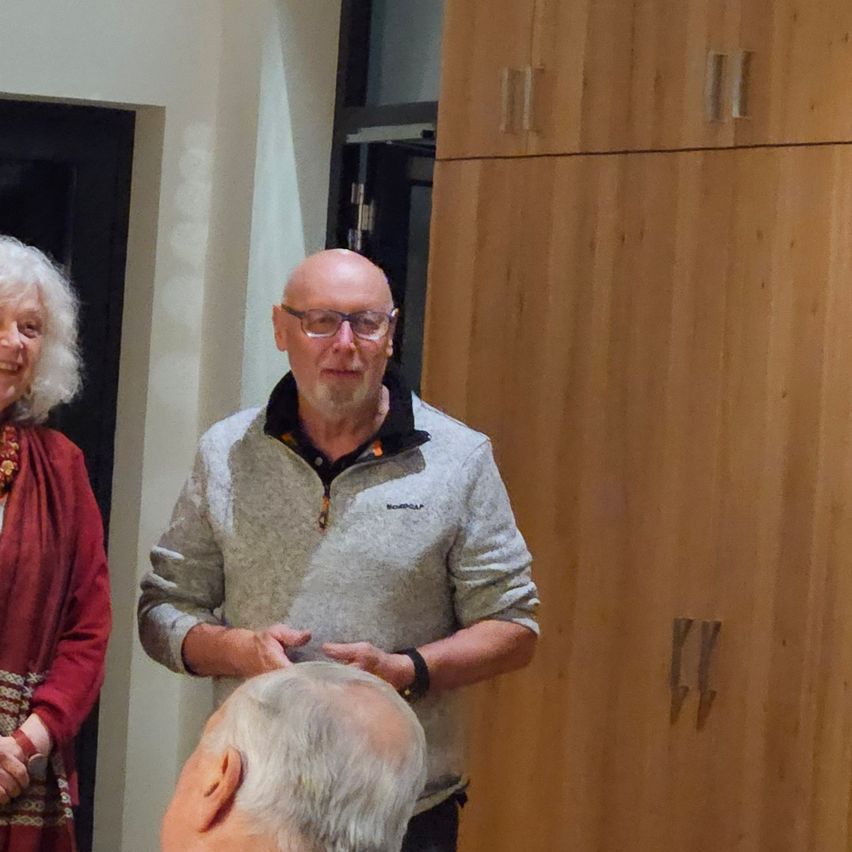 Ein Mann in einem grauen Pullover gestikuliert und spricht zu einer Frau in einem roten Kleid und einem Mann mit grauem Haar. Sie stehen vor einem Holzschrank.