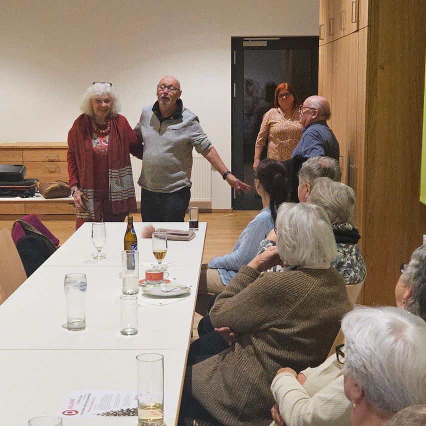 Eine Gruppe von Menschen sitzt an einem Tisch, einige stehen und andere sitzen. Ein Mann und eine Frau stehen in der Nähe der Tür, während andere in Gespräche vertieft sind. Weingläser und Teller stehen auf dem Tisch.