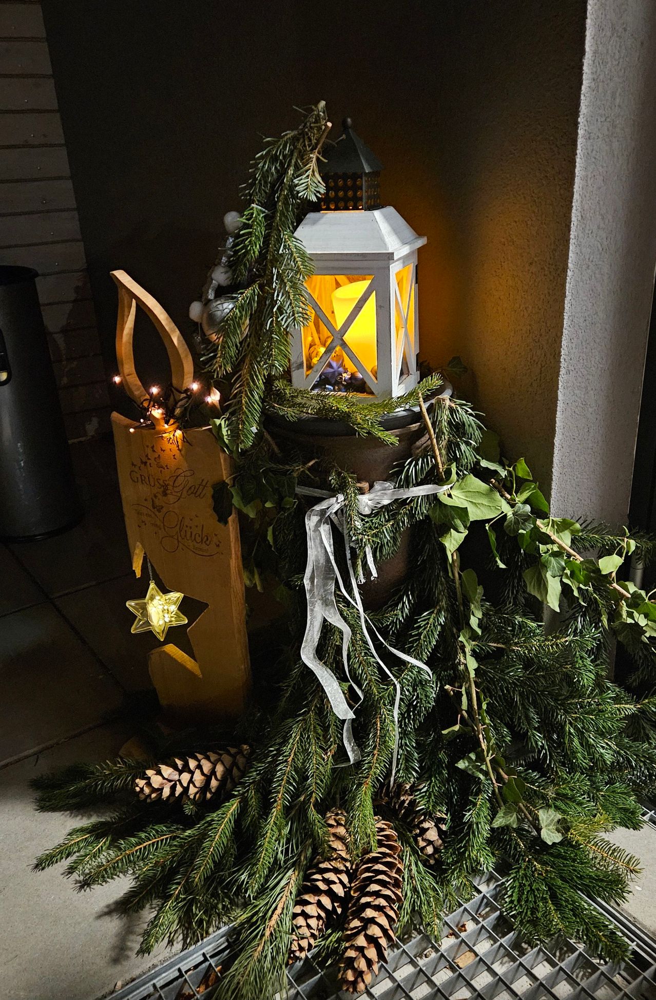 Eine Laterne mit einer Kerze ist in der Mitte eines Kranzes aus Tannenzweigen und Grün beleuchtet, dekoriert mit Lichtern und einem hölzernen Ornament.