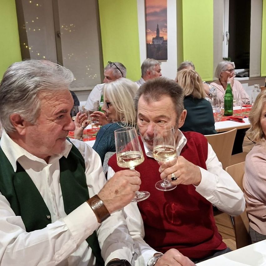 Eine Gruppe von Menschen sitzt an einem Tisch und stößt mit Weinläsern an. Zwei Männer sind im Vordergrund, mit einer grünen Flasche auf dem Tisch hinter ihnen.