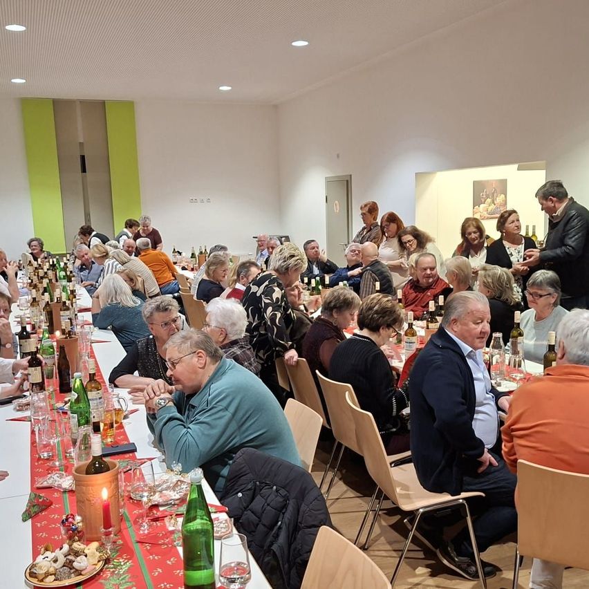 Viele Menschen sitzen um Tische in einem Saal. Die Tische sind mit rotem Tuch bedeckt, und Flaschen und Gläser sind darauf gestellt. Einige Leute stehen und unterhalten sich. An der Wand sind Bilderrahmen angebracht.