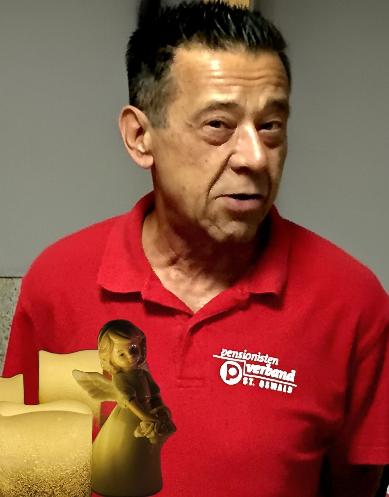 Ein älterer Mann in einem roten Hemd mit Logo und einer kleinen Engelsfigur auf der Brust.
