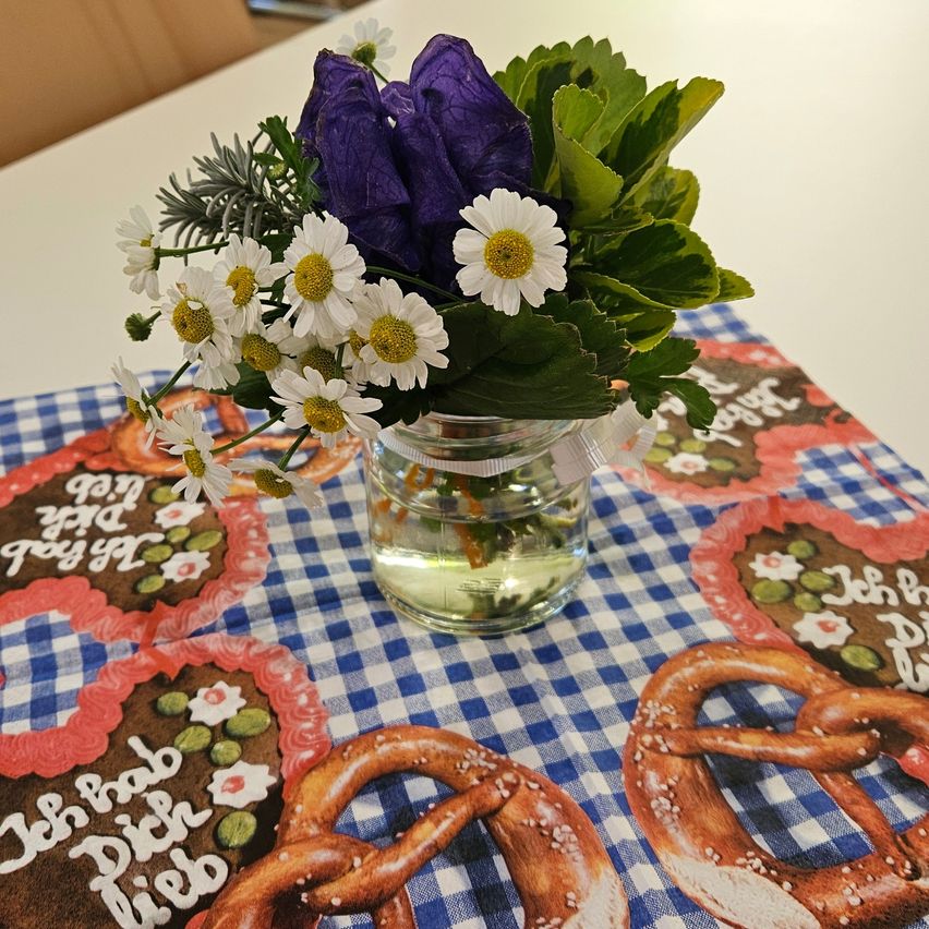Ein kleiner Strauß aus weißen Gänseblümchen und lila Blumen in einem Glas steht auf einem blau-weiß karierten Tuch, umgeben von Brezeln und herzförmigen Keksen.