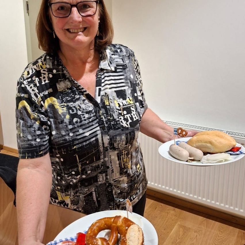Eine lächelnde Frau hält zwei Teller, einer mit Brötchen und einem Brezel, der andere mit verschiedenen Speisen, darunter Würstchen. Sie steht in einem Raum mit einer weißen Wand.
