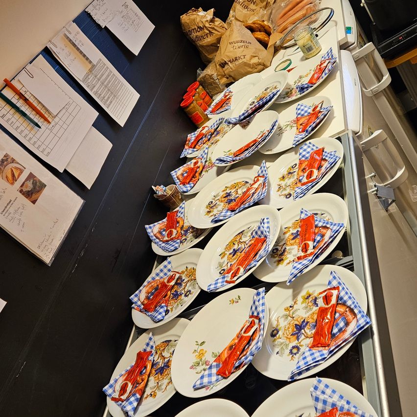 Eine Arbeitsplatte mit geordneten Tellern, jeder mit einem Stück Brot und einer Serviette. Dahinter befinden sich Lebensmitteltüten und Papiere.