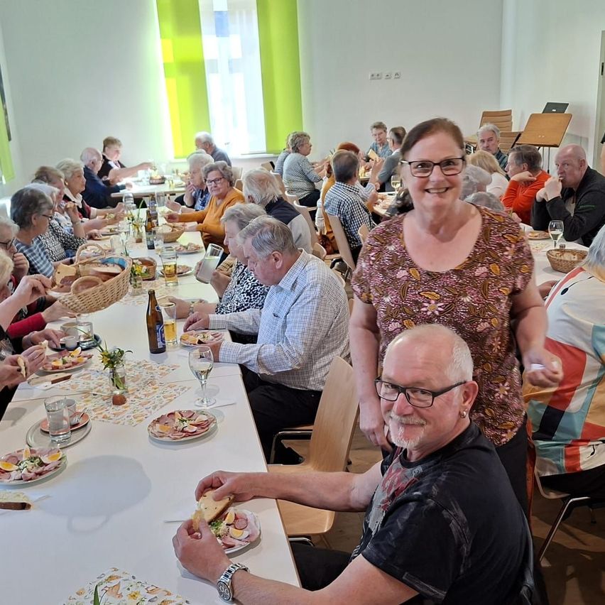 A group of people sit at a long table in a dining hall, smiling and posing for a photo. They are surrounded by plates, wine glasses, and a basket of bread.