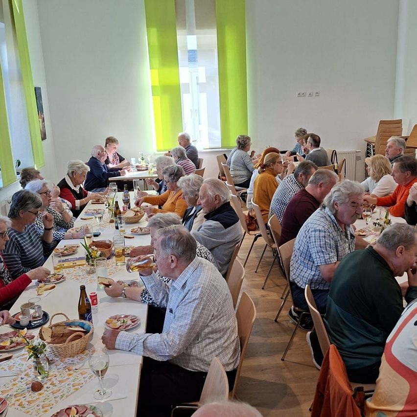 A group of older adults are seated at a long table, eating and drinking. The table is filled with food and drinks. There are chairs on both sides of the table. In the background, there is a window with a view of the outside.