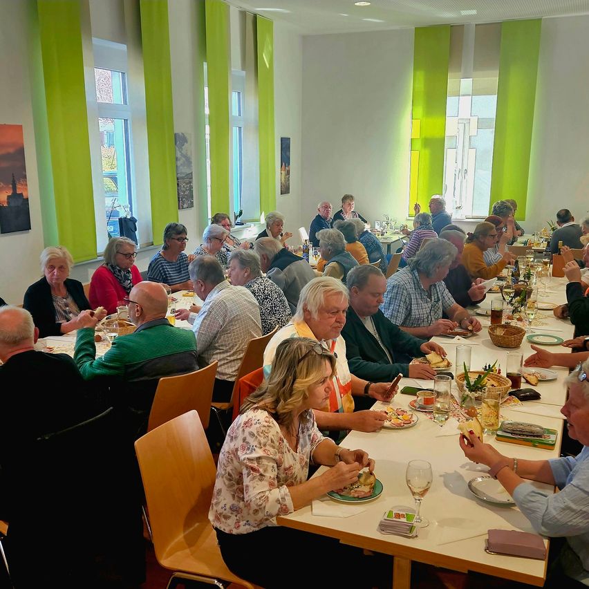 A group of people are seated at a long table, possibly in a restaurant, enjoying their meal. The table is set with plates, wine glasses, and other items. The room has white walls and green curtains.
