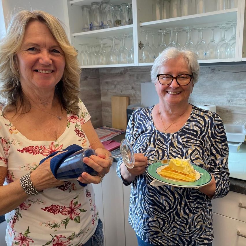 Two women stand in a kitchen, one holding a bottle and the other a plate with a lemon dessert. They are smiling and appear to be posing for a photo.