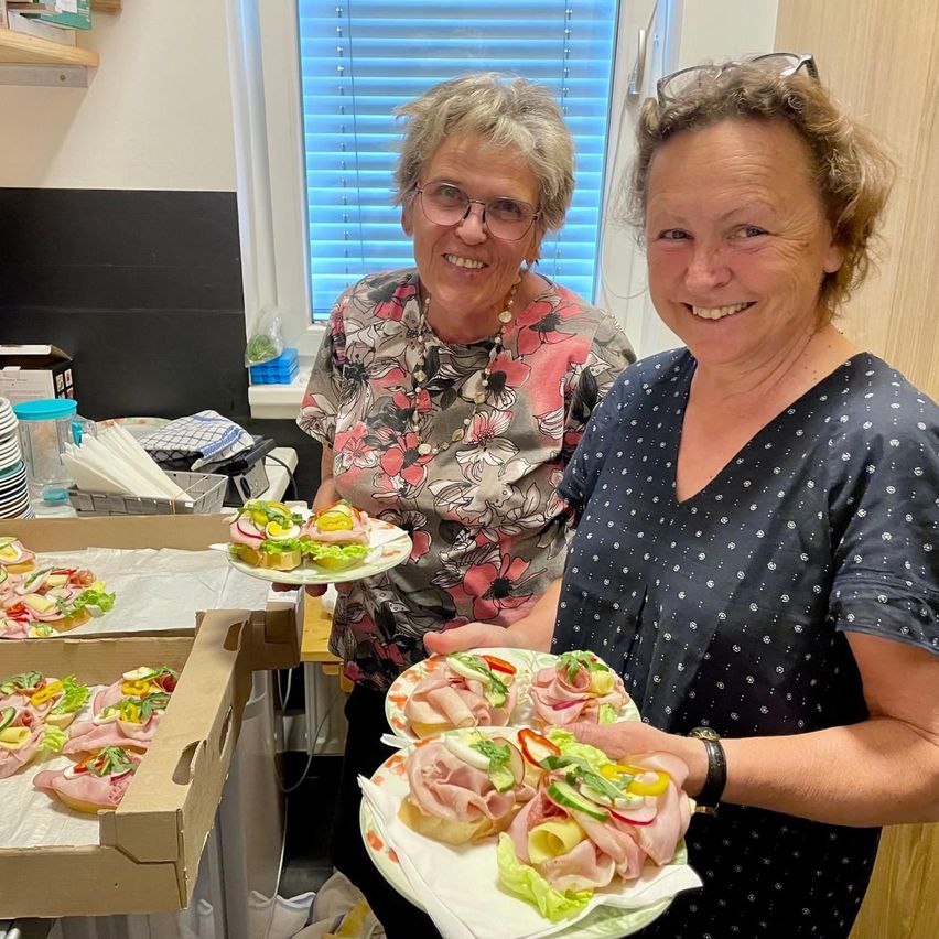 Two women holding plates with sandwiches in a kitchen, smiling for a photo. One woman wears glasses and a necklace.