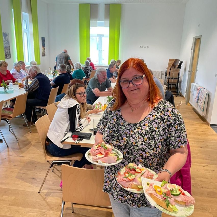 A woman with glasses and red hair holds plates of sandwiches in a dining room. People sit at tables, and a woman in white sits on a chair.