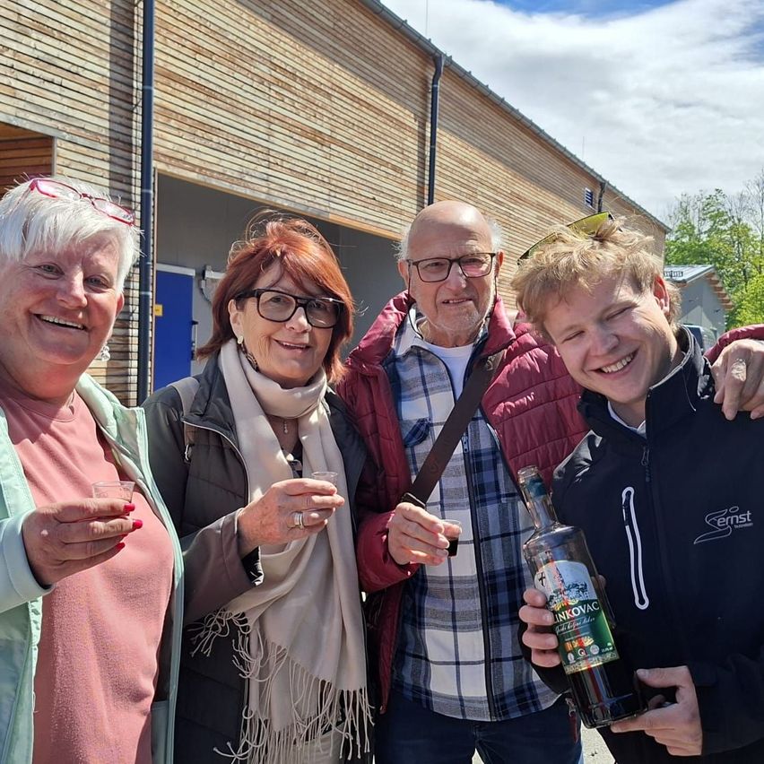 Eine Gruppe von vier Personen lächelt und posiert für ein Foto draußen. Zwei Frauen und zwei Männer. Der Mann auf der rechten Seite hält eine Flasche.
