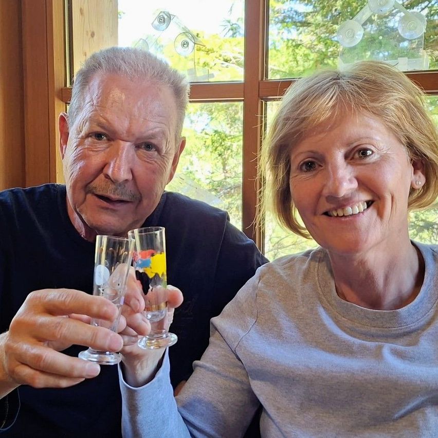 Ein älterer Mann und eine ältere Frau lächeln und halten zwei Gläser in den Händen. Sie sitzen nebeneinander mit einem Fenster dahinter.