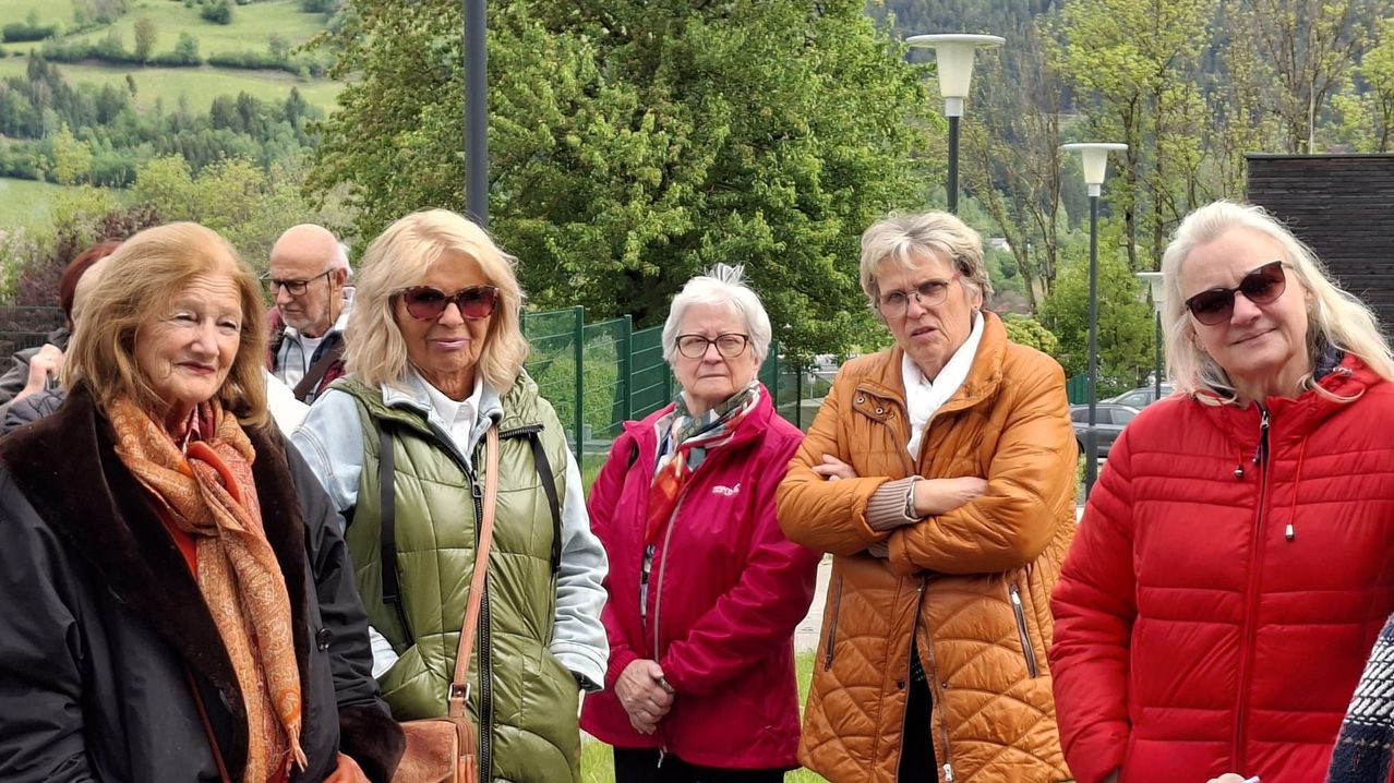 Eine Gruppe älterer Frauen steht im Freien, lächelt und posiert für ein Foto in einem Park. Hinter ihnen sind Bäume und ein Zaun zu sehen, mit Bergen in der Ferne.