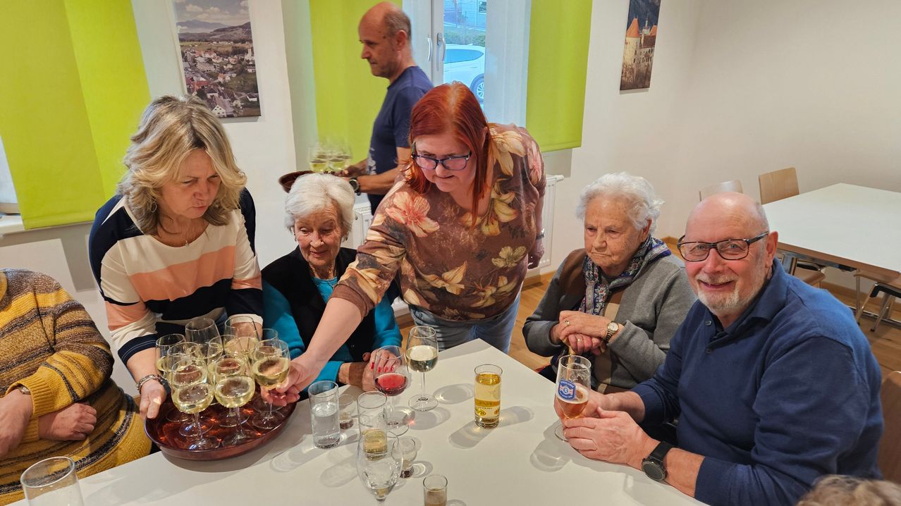 A group of people, including elderly women and men, are seated around a table, enjoying drinks. The woman with red hair pours a drink, while another woman holds a glass.