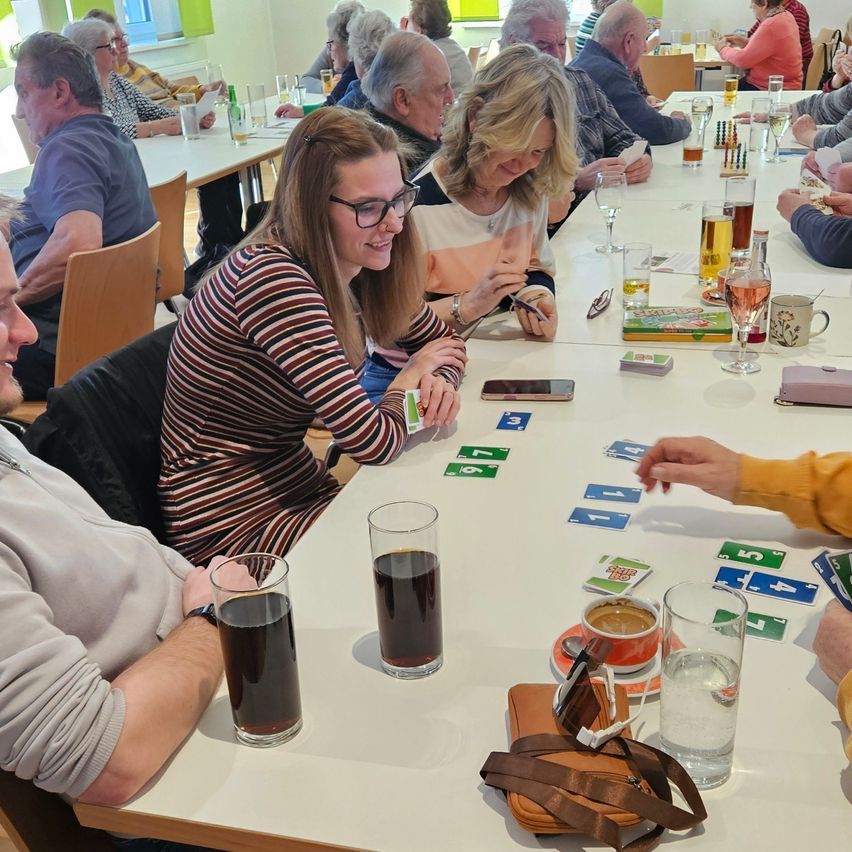 A group of elderly people are sitting around a table in a room, playing a card game. Some are holding glasses, and some have food on their plates.
