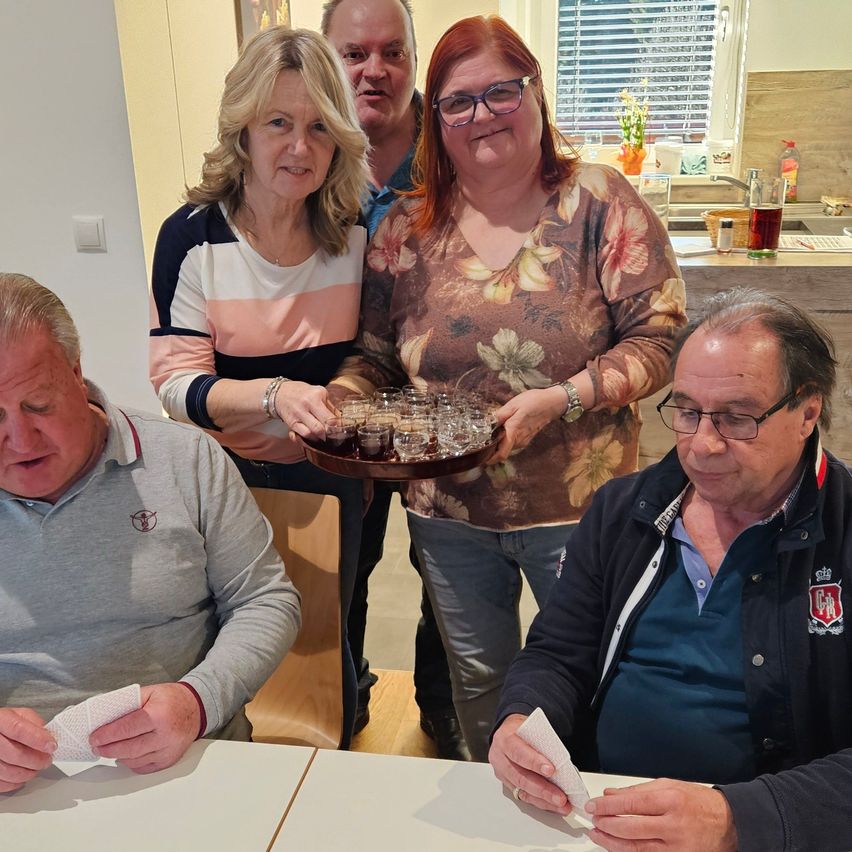 Five adults sit at a table, two women stand behind with drinks. One woman holds a tray of small glasses. The room has wooden floors and a kitchen in the background.